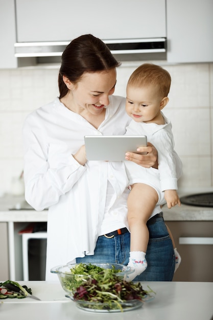 Mulher em pé na cozinha com o bebê nas mãos, mostrando algo na tela do tablet digital