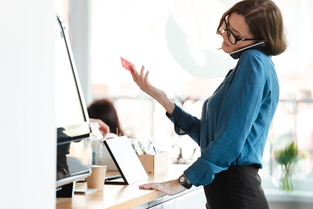 Mulher em pé no café, segurando o cartão de crédito, falando por telefone