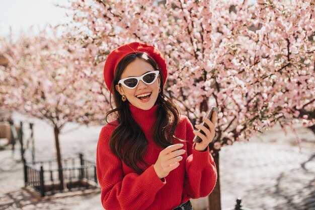 Mulher em poses de alto astral com telefone perto das flores de cerejeira. Retrato de senhora de boina vermelha, suéter cashemere e óculos brancos no jardim na primavera