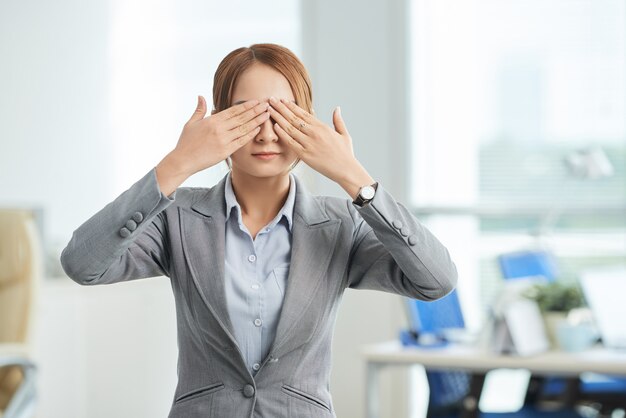 Mulher em um terno de negócio em pé no escritório com as mãos cobrindo os olhos