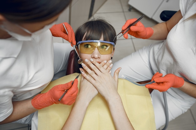 Mulher em uma cadeira odontológica. A menina cobre a boca. Os dentistas tratam os dentes de uma menina