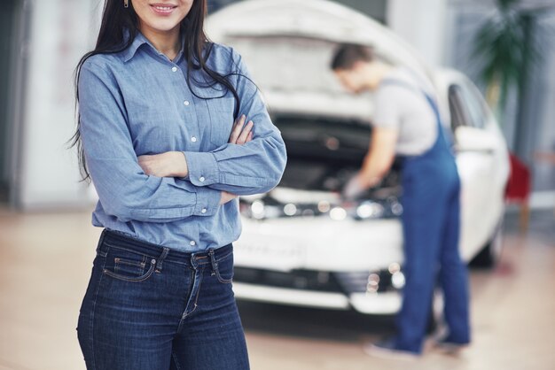 Mulher em uma garagem de carro, recebendo serviço mecânico. O mecânico trabalha sob o capô do carro