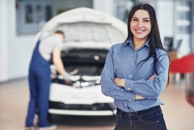 Mulher em uma garagem de carro, recebendo serviço mecânico. O mecânico trabalha sob o capô do carro