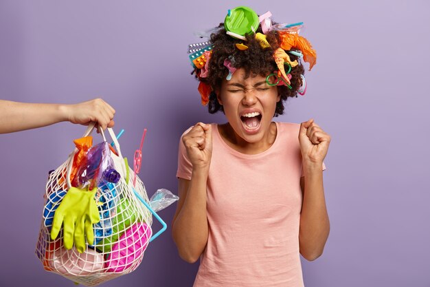 Mulher emocionalmente indignada, ativista ambientalista ergue os punhos cerrados, grita alto, tem olhar deprimido, grita alto para não poluir nossa Terra cercada de muito lixo plástico vestido casualmente.