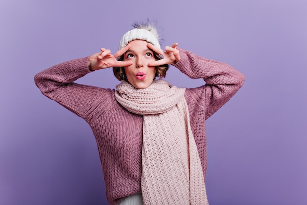 Mulher emocionalmente satisfeita num cachecol de malha quente, brincando e fazendo caretas. linda garota caucasiana com chapéu e suéter grande isolado na parede brilhante.