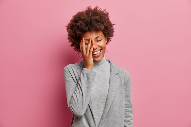 Mulher empresária de pele escura radiante ri positivamente, fica de olhos fechados, ri de piada engraçada, faz cara de palma, vestida com elegância, mostra dentes brancos