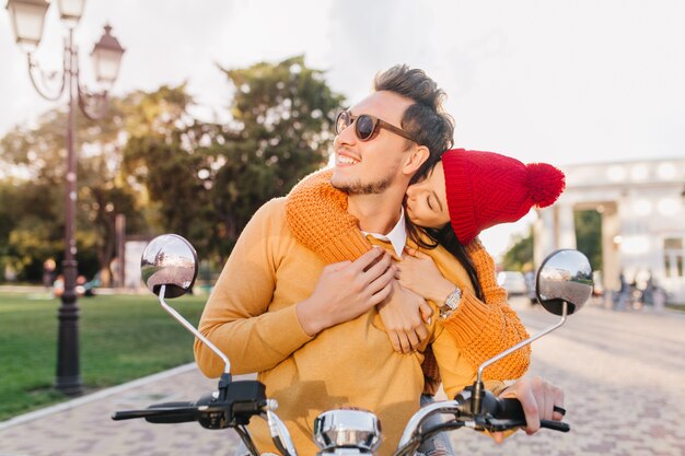 Mulher encantadora com chapéu vermelho beijando o pescoço do namorado sentada na scooter