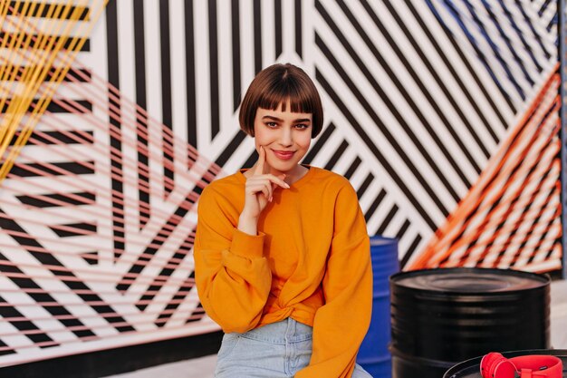 Mulher encantadora com penteado curto fofo sorrindo em pano de fundo listrado Linda garota de moletom laranja e jeans posando ao ar livre