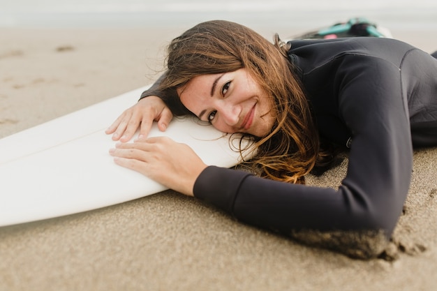 Mulher encantadora com roupa de neoprene deitada na prancha de surf na praia em um dia ensolarado e quente