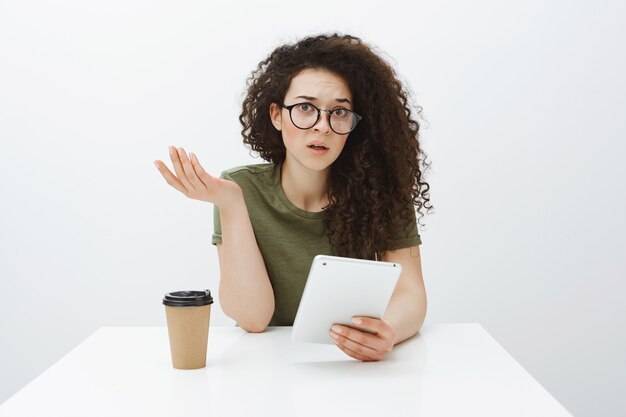 Mulher encantadora confusa sem noção com cabelo encaracolado nos óculos, levantando a mão em um gesto questionador, sentada à mesa