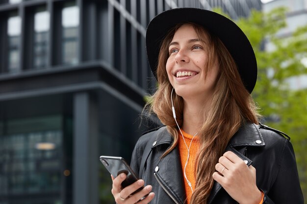 Mulher encantadora e feliz acessa o site com música moderna, segura o celular, usa fones de ouvido e usa chapéu preto