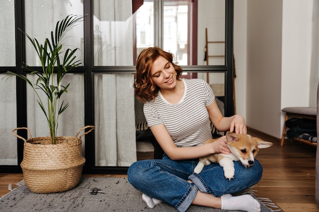 Foto grátis mulher encantadora em jeans está descansando na sala de estar e brincando com o cachorro.