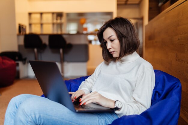 Mulher encantadora está sentado na cadeira de bolsa azul, trabalhando no laptop, falando no telefone