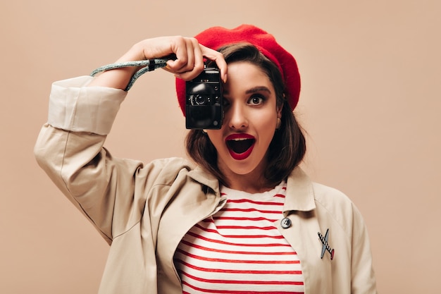 Foto grátis mulher encantadora na boina e trincheira tira foto no fundo isolado. maravilhosa jovem com lábios vermelhos e suéter listrado está segurando a câmera.