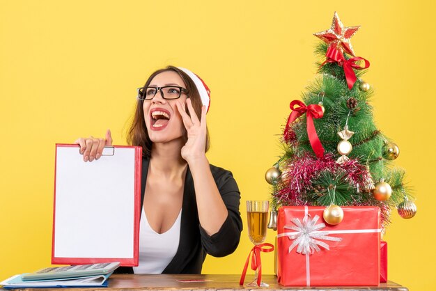 Mulher encantadora sorridente de terno com chapéu de Papai Noel e óculos mostrando documento chamando alguém no escritório em amarelo isolado