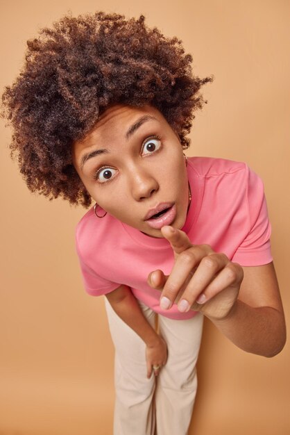 mulher encara os olhos esbugalhados aponta diretamente para a câmera percebe algo chocante vestida casualmente isolada em bege. Nossa, que coisa impressionante