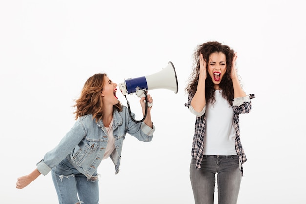 Foto grátis mulher encaracolada confusa, cobrindo as orelhas enquanto segunda garota gritando com ela com megafone sobre parede branca