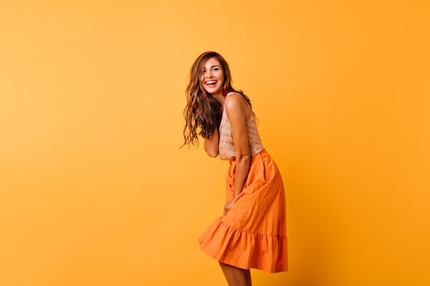 Foto grátis mulher encaracolada de cabelos compridos em roupa brilhante, expressando emoções positivas. modelo feminino romântico em saia laranja dançando em amarelo.