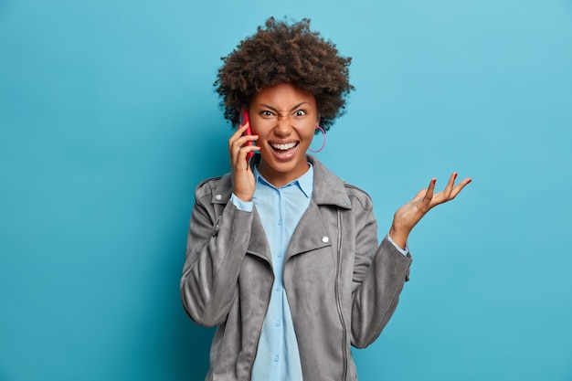 Foto grátis mulher encaracolada de pele escura irritada e irritada levanta a mão, conversa ao telefone, veste roupas elegantes, faz gestos ativamente