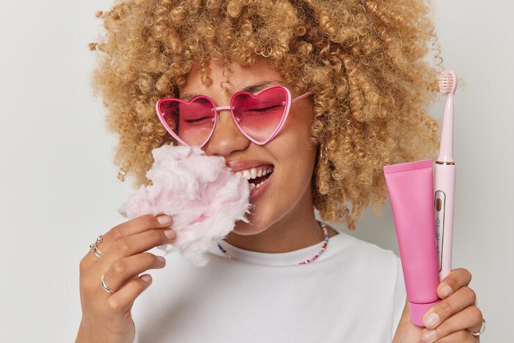 Foto grátis mulher encaracolada feliz come algodão doce saboroso consome comida prejudicial para os dentes segura escova de dentes elétrica e tubo de pasta de dente usa óculos de sol rosa e camiseta casual isolada sobre fundo branco
