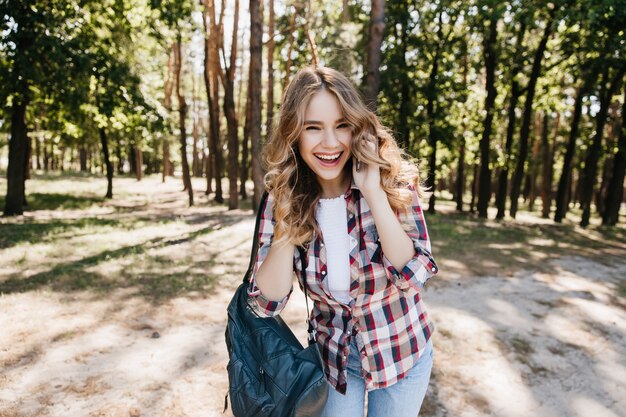 Mulher encaracolada feliz posando com o telefone no parque de verão e rindo. Menina caucasiana bem vestida que expressa emoções positivas sinceras.