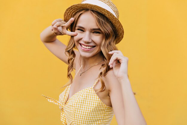 Mulher encaracolada levemente bronzeada com chapéu sorrindo. Foto interna de uma modelo feminina inspirada usando um traje amarelo xadrez.