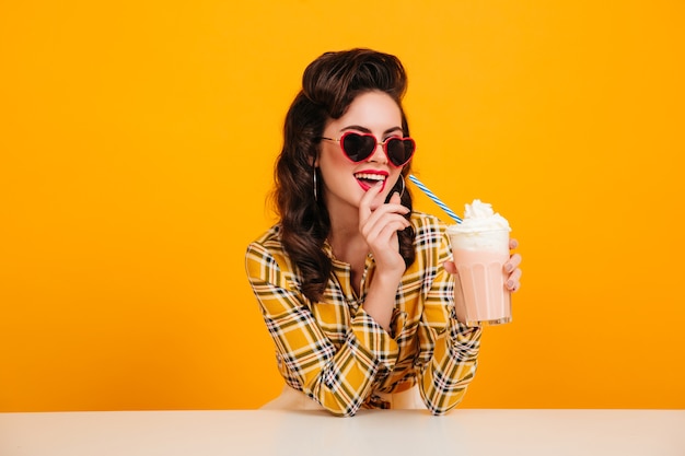 Mulher encaracolada maravilhosa desfrutando de milk-shake. Foto de estúdio de garota Pin-up com coquetel posando em fundo amarelo.