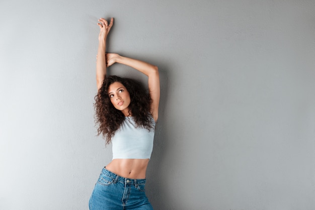 Foto grátis mulher encaracolada muito sorridente posando no estúdio com as mãos acima da parede cinza e olhando para cima