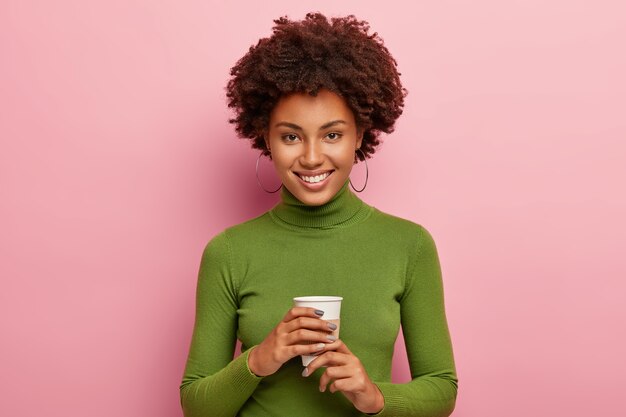Mulher encaracolada satisfeita aprecia a pausa para o café, segura um copo descartável de bebida, parece feliz, usa gola olímpica verde, sorri com alegria, tem tempo livre depois do trabalho isolado na parede rosa