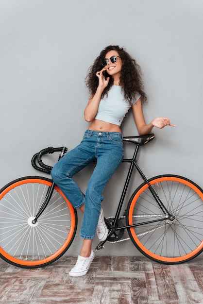 Mulher encaracolada sorridente em óculos de sol posando com bicicleta e falando pelo smartphone