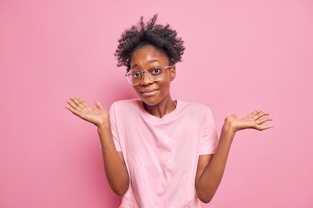 Foto grátis mulher encolhe os ombros mostra as mãos vazias não tem idéia de algo nada a dizer fica inconsciente interior usa grandes óculos ópticos camiseta casual isolada na parede rosa