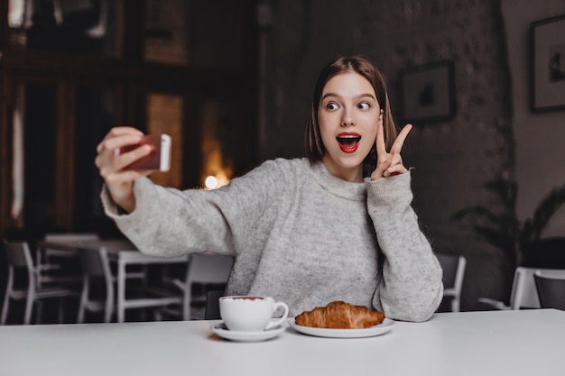 Mulher enérgica de suéter cinza e batom vermelho faz selfie. retrato de menina mostrando sinal de paz no café com croissant na mesa.