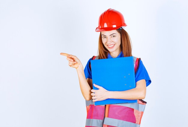 Mulher engenheira de capacete vermelho segurando uma pasta de projeto azul e apresentando alguém ou algo.