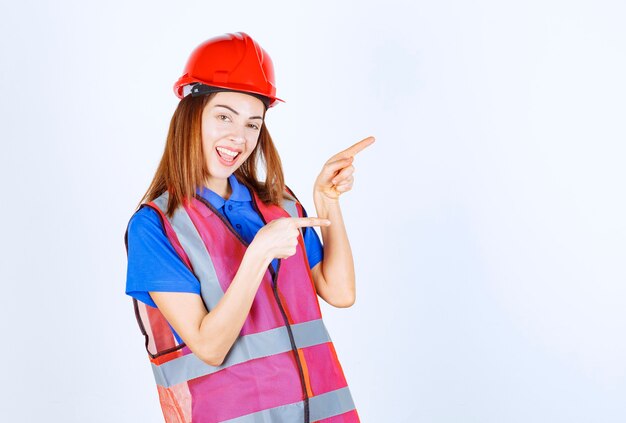 Foto grátis mulher engenheira de uniforme e capacete vermelho apontando para alguém à direita.