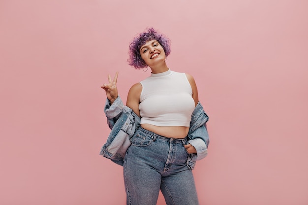 Mulher engraçada encaracolada com cabelo lilás com um sorriso adorável na camiseta branca mostra sinais de paz em rosa.
