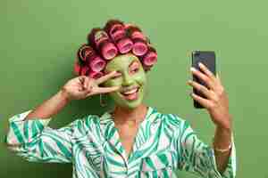 Foto grátis mulher engraçada feliz faz selfie formas sinal de vitória na câmera do smartphone sorri amplamente aprecia tratamentos faciais; aplique rolos de cabelo vestidos com roupas casuais domésticas, isolados sobre a parede verde