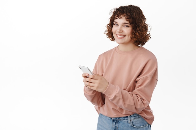 mulher enviando mensagens de texto, usando o aplicativo móvel do smartphone, sorrindo feliz para a câmera, mensagens, bate-papo no aplicativo, de pé no branco.