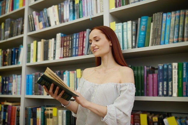 Mulher envolvida lendo livro em pé perto de estantes
