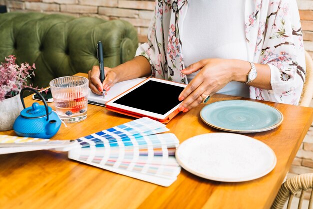 Mulher escrevendo no notebook com tablet digital; Placas e amostra de cor na mesa de madeira