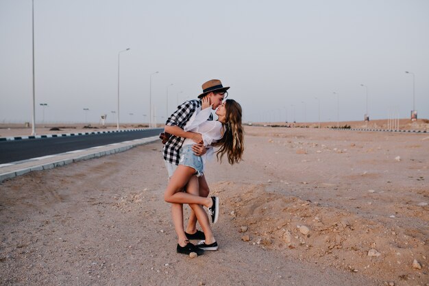 Mulher esguia em shorts jeans em uma perna, beija suavemente o namorado no lindo deserto. Jovem elegante abraçando a namorada, posando perto da rodovia nas férias de verão