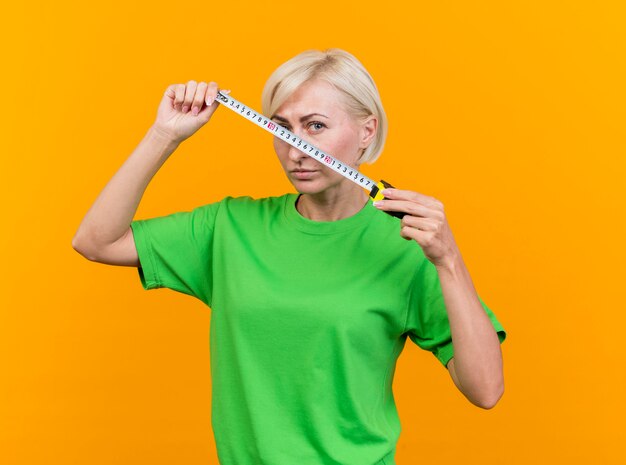 Foto grátis mulher eslava loira confiante de meia-idade olhando para a câmera segurando o medidor de fita na frente do rosto isolado em um fundo amarelo