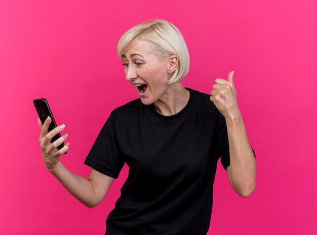 Mulher eslava loira de meia-idade alegre segurando e olhando para o celular, mostrando o polegar isolado na parede rosa