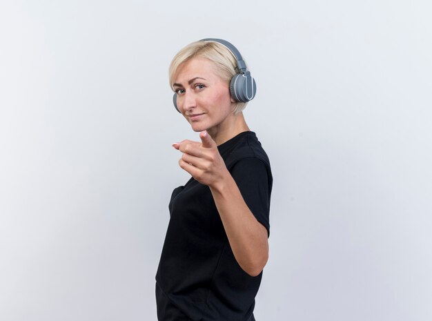 Mulher eslava loira de meia-idade confiante usando fones de ouvido em vista de perfil, olhando e apontando para a câmera, isolada no fundo branco com espaço de cópia