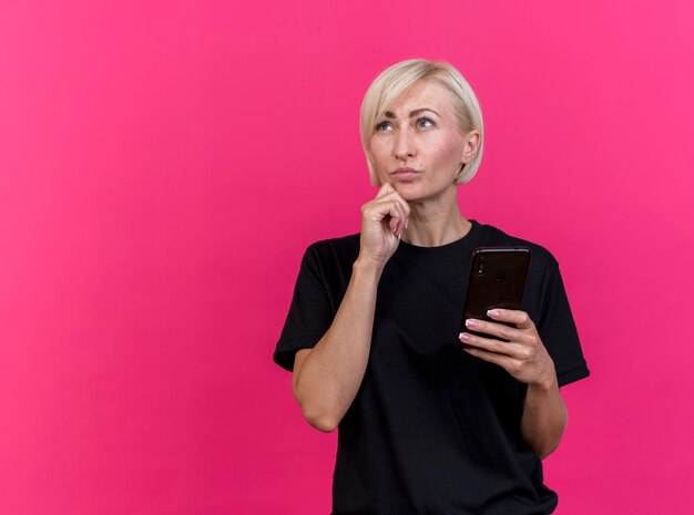 Mulher eslava loira de meia-idade pensativa segurando um telefone celular, tocando o queixo, olhando para cima, isolada em um fundo carmesim com espaço de cópia