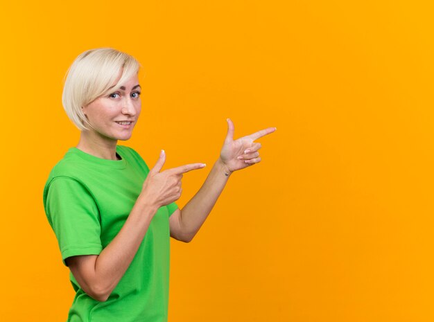 Mulher eslava loira de meia-idade sorridente em pé em vista de perfil, olhando para a câmera apontando diretamente, isolada em um fundo amarelo com espaço de cópia