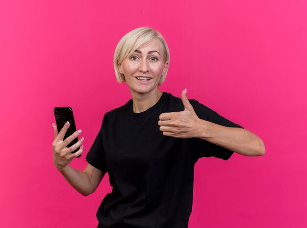 Mulher eslava loira de meia-idade sorridente segurando um celular, olhando para a frente, mostrando o polegar isolado na parede rosa