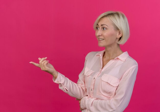 Mulher eslava loira sorridente em pé em vista de perfil, olhando e apontando diretamente, isolado em um fundo rosa