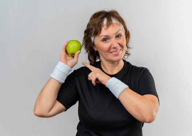 Mulher esportiva de meia-idade em uma camiseta preta com fita na cabeça segurando duas maçãs verdes apontando com o dedo para elas sorrindo alegremente em pé sobre uma parede branca