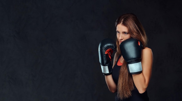 Mulher esportiva vestida com roupas esportivas usando luvas de boxe posando em um estúdio. isolado em plano de fundo texturizado escuro.