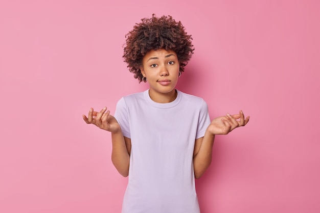 mulher estende as palmas das mãos para os lados tem expressão duvidosa expressa incerteza usa camiseta casual isoturada em suportes rosa sem saber nada sabe